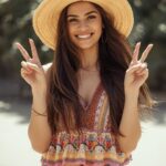 lange braune haare frau mit braunen augen hält 2 finger hoch. und zeigt damit die nummer 2 an. sie trägt kurze shorts und ein cooles boho sommeroberteil. mit einem coolen hut.